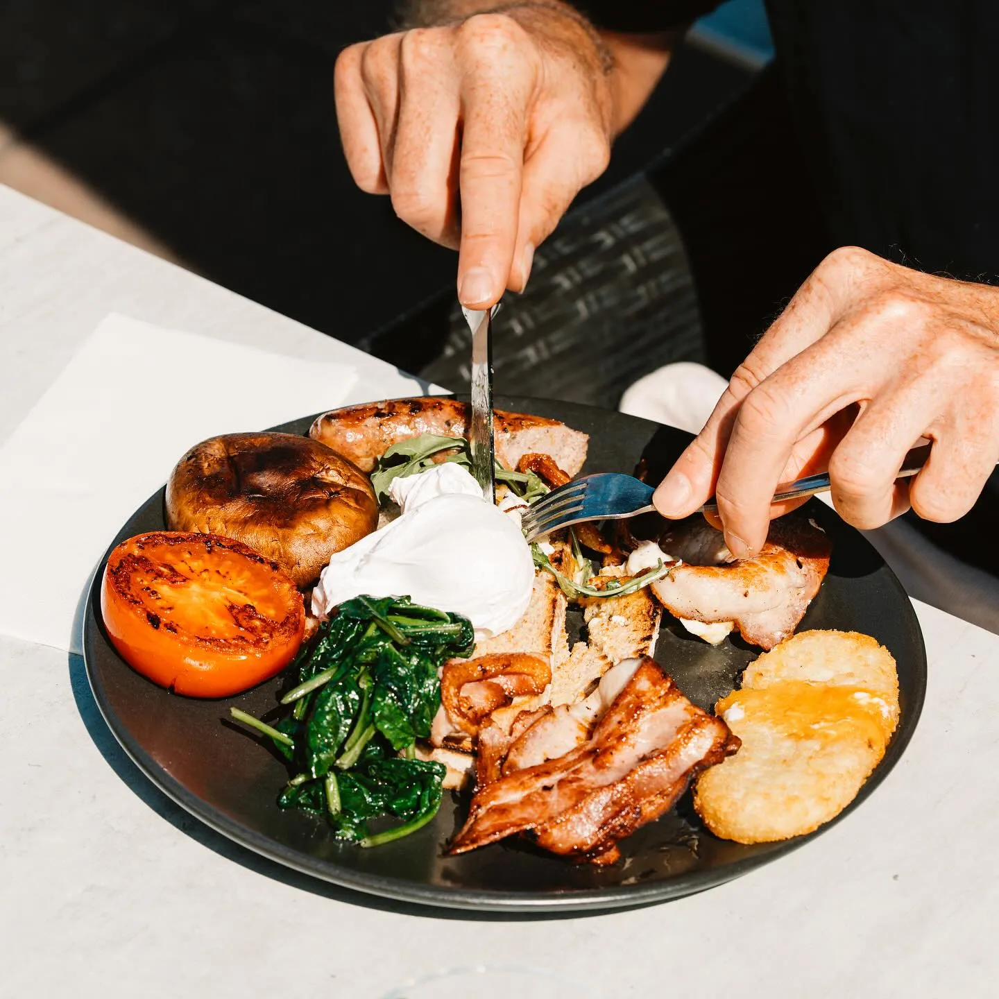Hands cutting into a big breakfast with poached egg, bacon, sausage, mushrooms, spinach, grilled tomato and hash brown
