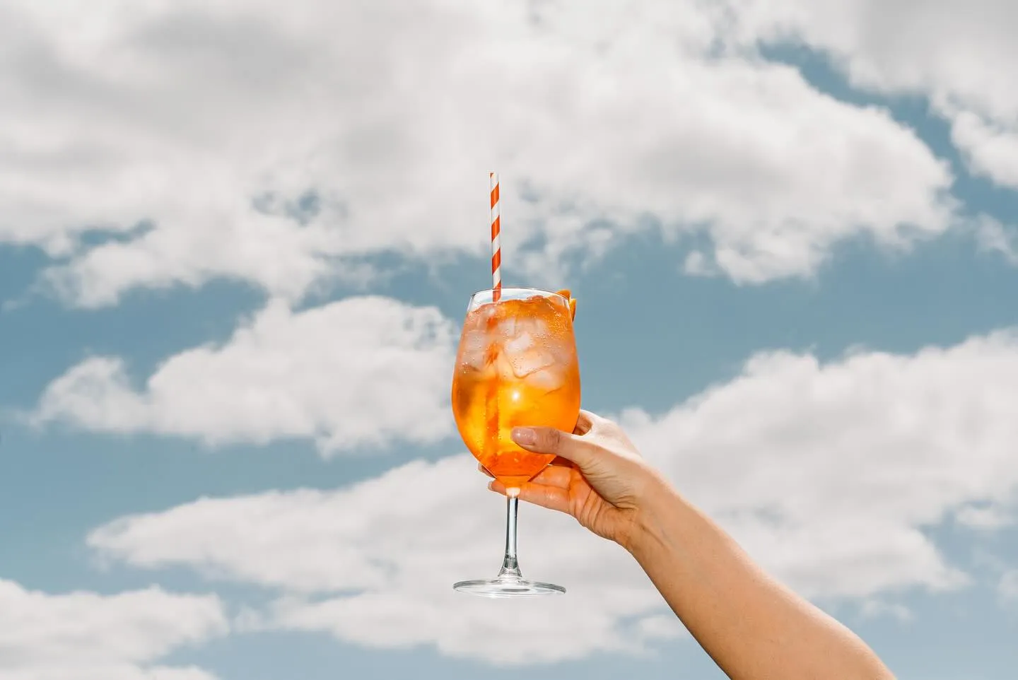 Hand holding an Aperol Spritz with striped straw against a cloudy sky